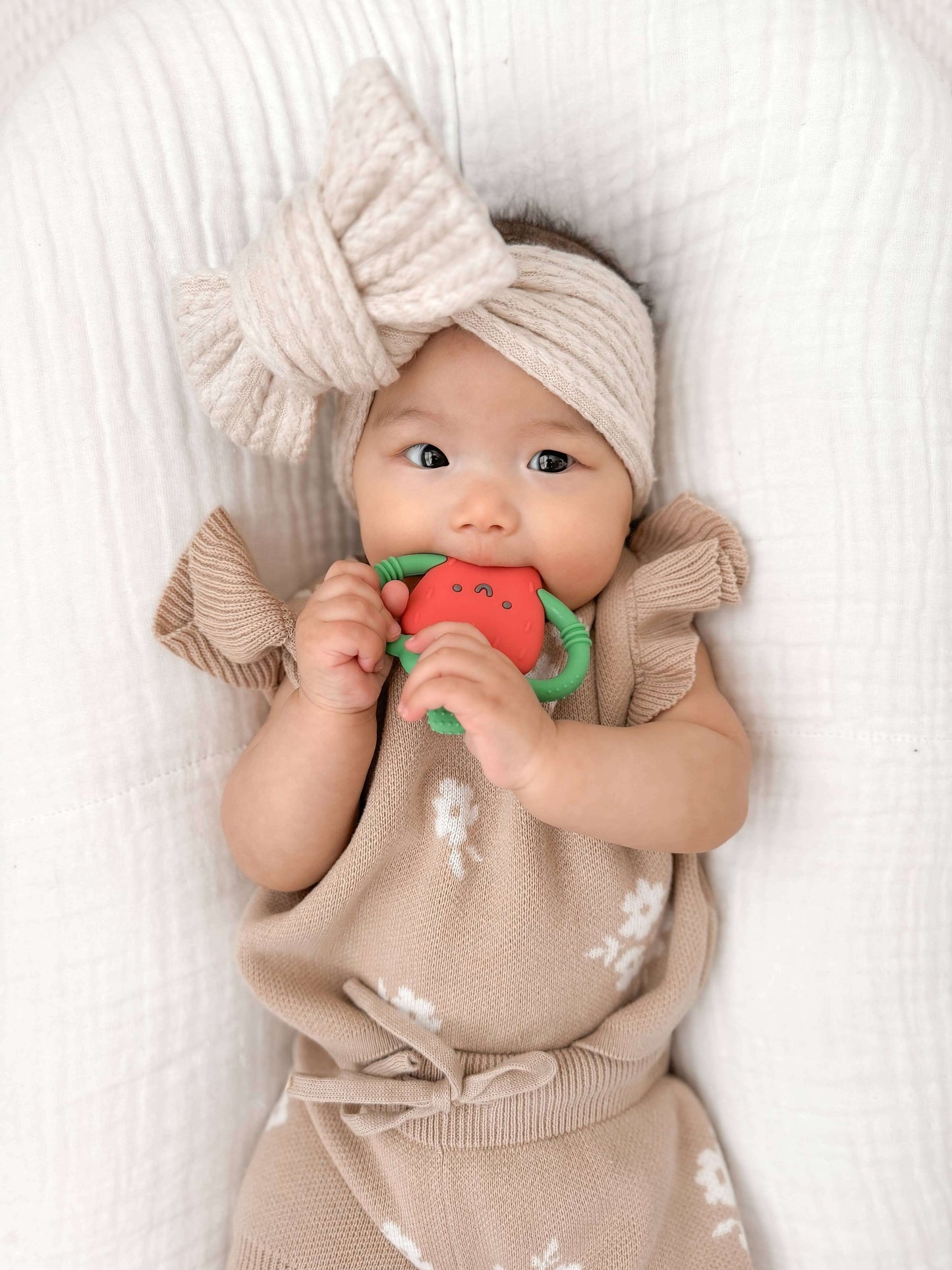Baby holding Bonnie the Strawberry teether from the Chew Crew collection by Itzy Ritzy, wearing a beige outfit and knit headband