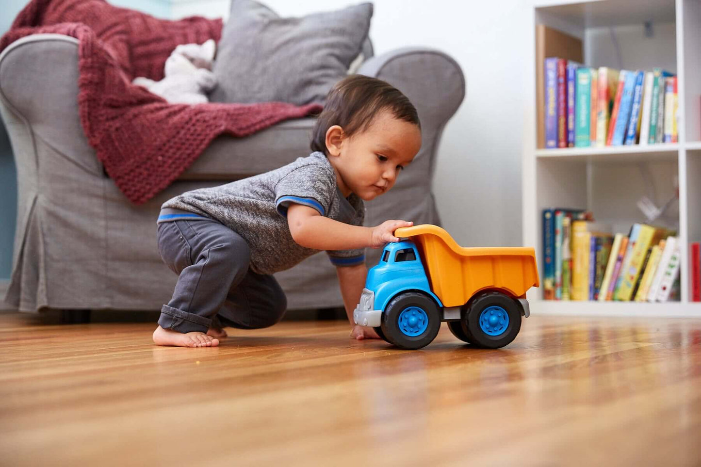 Toddler playing with eco-friendly blue and orange dump truck made from recycled plastic on wooden floor.