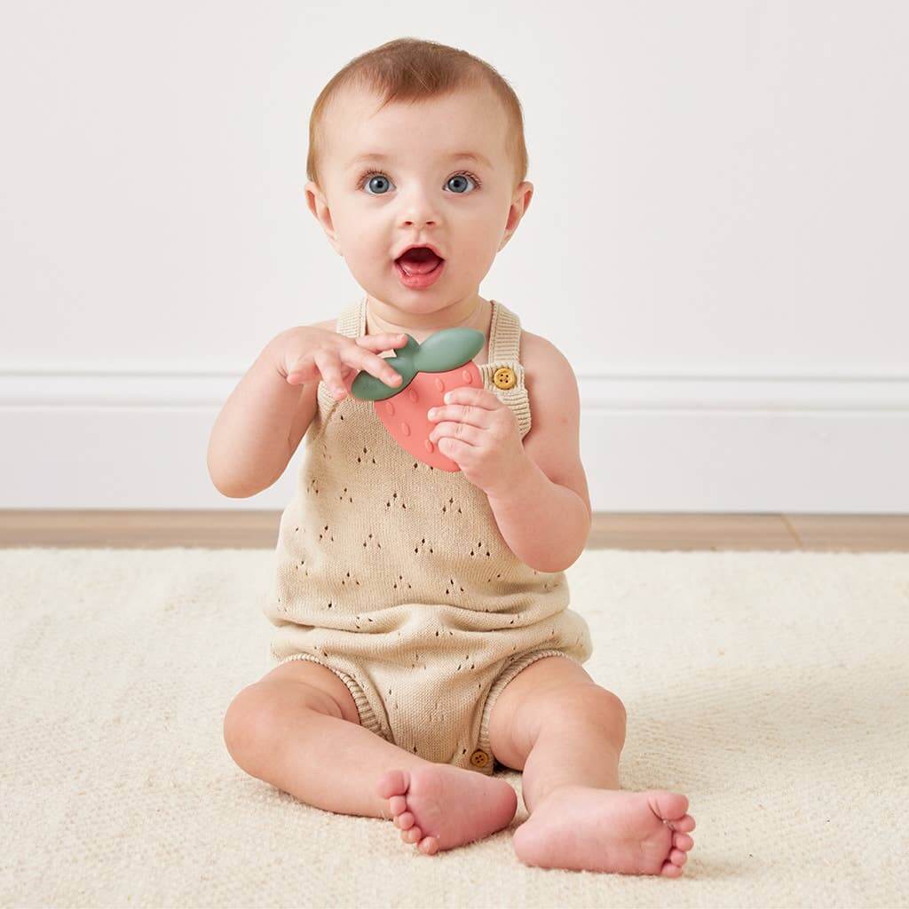 Baby holding Strawberry Sweetie Rattle™ silicone teether, sitting on a cream-colored carpet, showing excitement.